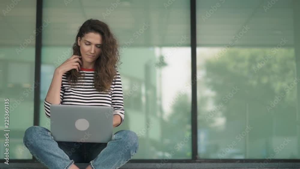 Serious young woman with long curly hair is sitting with crossed legs on building stairs and typing at her laptop keyboard. Handheld real time medium shot Serious young woman with long curly hair is sitting with crossed legs on building stairs and typing at her laptop keyboard. Handheld real time medium shot