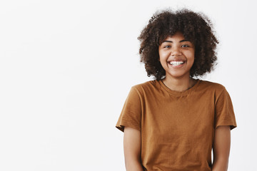 Waist-up shot of cute carefree friendly-looking African American teenage girl with afro hairstyle smiling broadly with shy and happy expression meeting new classmates over gray background
