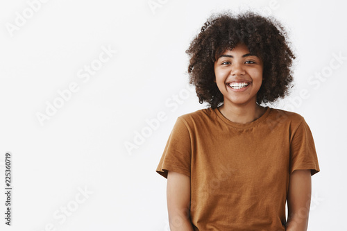 Waist-up shot of cute carefree friendly-looking African American teenage girl with afro hairstyle smiling broadly with shy and happy expression meeting new classmates over gray background