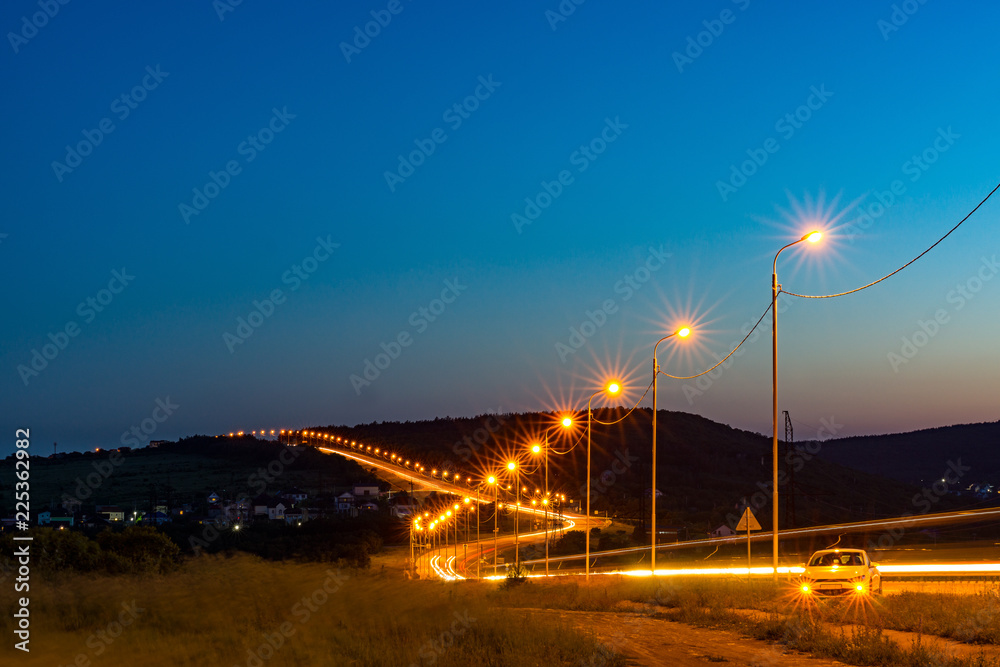 The night road meanders away into the distance. It is lit by the lights of passing cars and street lamps.