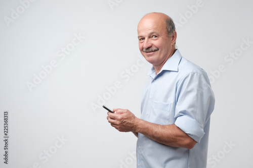 Mature businessman with mustache in blue shirt using phone to write sms. Isolated on gray background