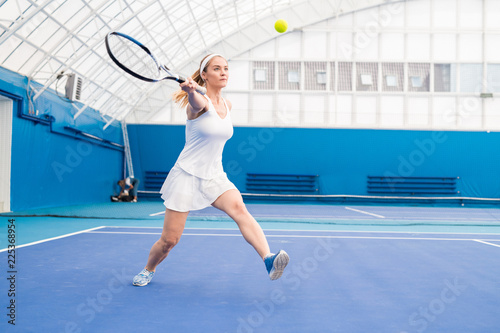 Full length portrait of female tennis player hitting ball during training in indoor court, copy space