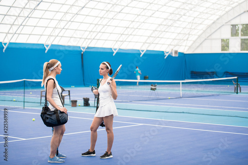 Full length portrait of two young women chatting in indoor tennis court after practice, copy space