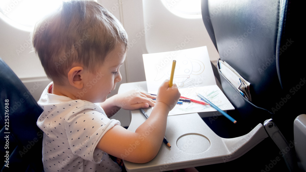 Naklejka premium Toned portrait of little toddler boy traveling by plane and drawing with pencils during long flight