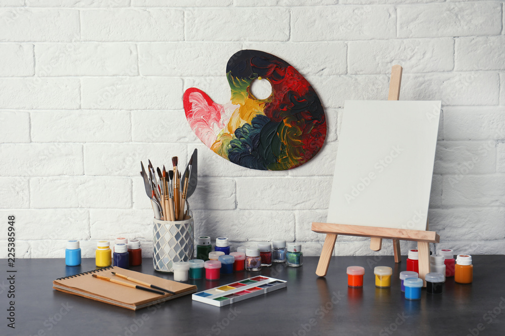 Naklejka premium Easel with various artist tools on table against brick wall. Space for text