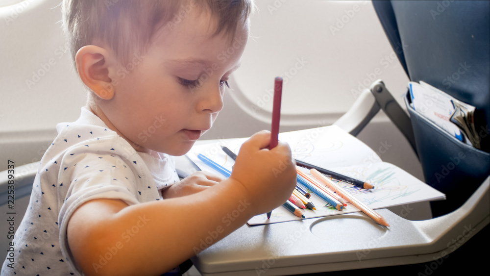 Closeup portrait of little toddler boy drawing with colorful pancils on foldable table in airplane during long flight