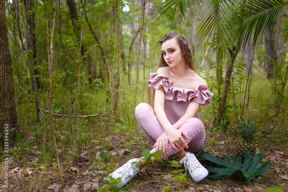 a young teenager girl with long curled hair dressed in pink sitting in the forest with a pineapple and exotic plant leaf