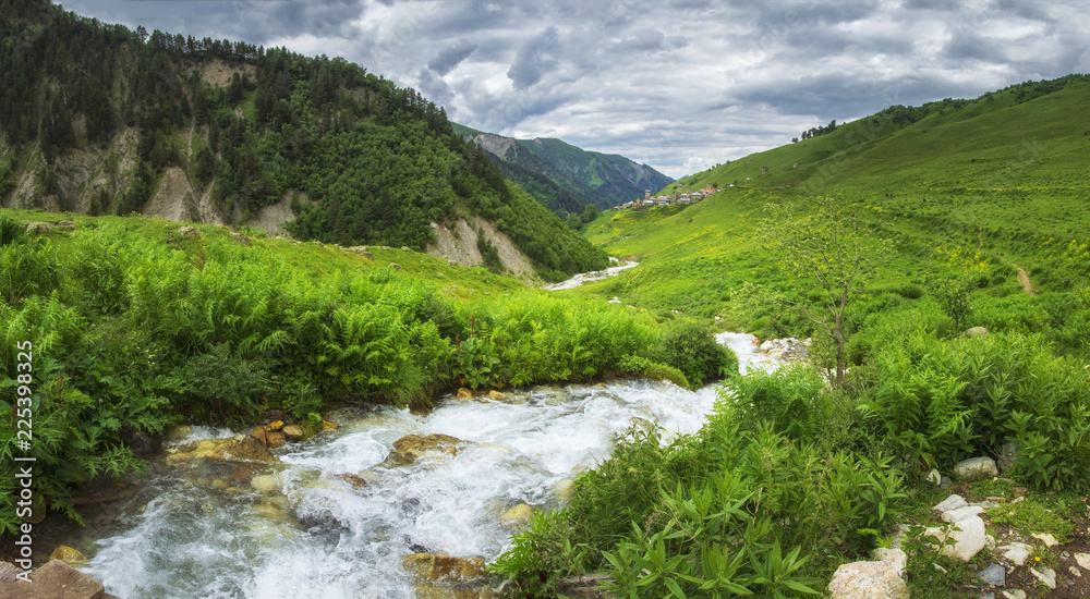 Georgian nature landscape in Svaneti region with mountain river in highlands. Green hills and mountains covered by grass. Cloudy sky over village Adishi on horizon, Georgia.