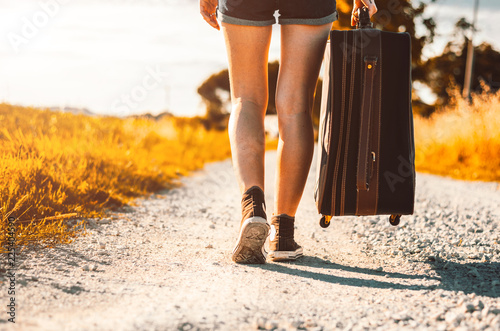 Close up of legs of a woman at vacation walking on the road holding her suitcase on a countryside background photo
