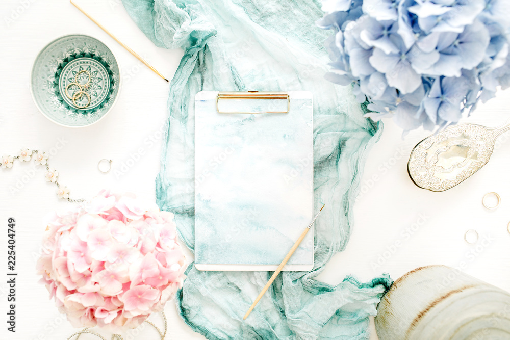 Artist home office desk workspace with watercolor clipboard, paintbrush, turquoise blanket, colorful pastel hydrangea flower bouquet, woman fashion accessories on white background. Flat lay.