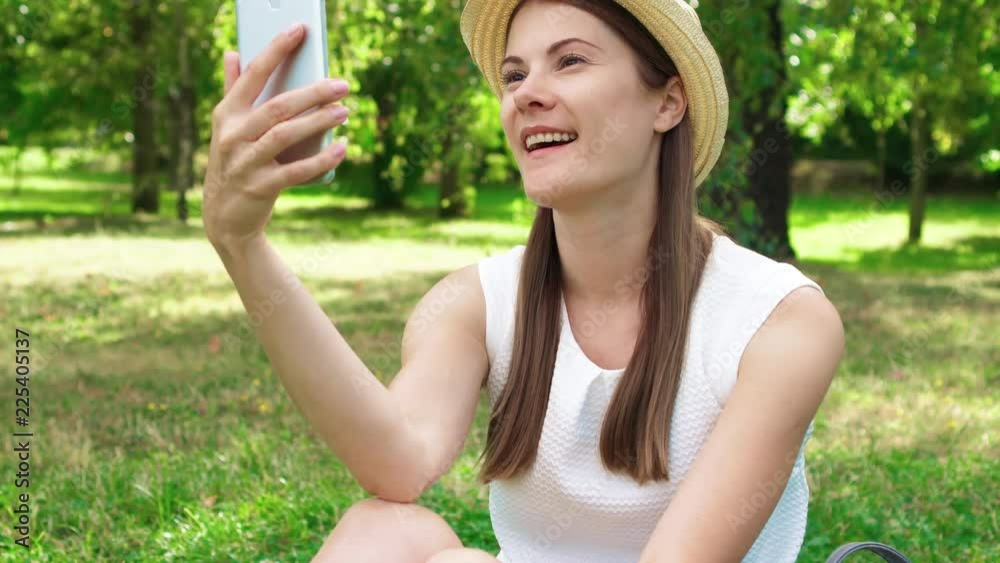 Young female student in white shirt sitting on grass on college campus using mobile phone. Tourist in hat having video chat via online app on cellphone in public park enjoying summer sunny day Young female student in white shirt sitting on grass on college campus using mobile phone. Tourist in hat having video chat via online app on cellphone in public park enjoying summer sunny day