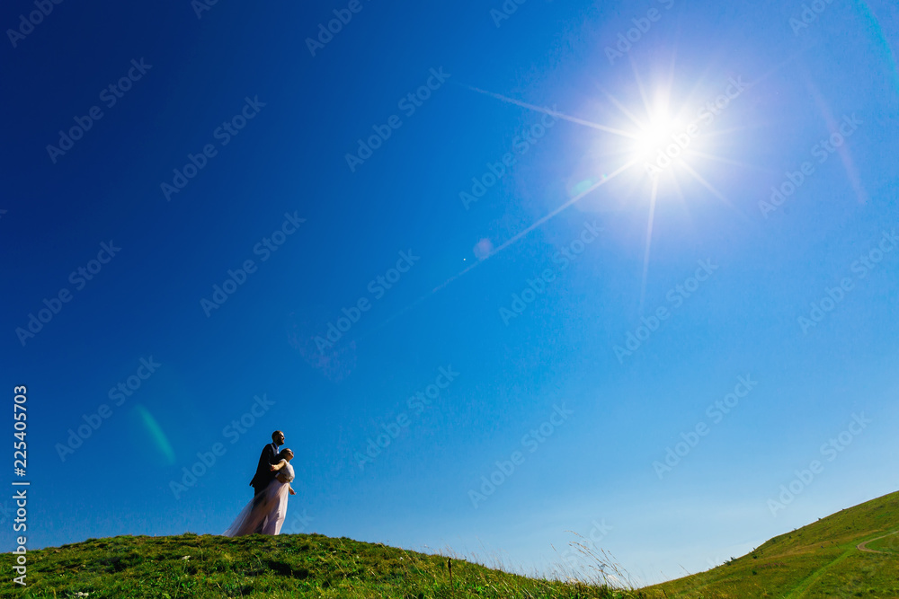 the groom hugging his bride from the back side behind her waist on top of a mountain with green grass on the background of blue sky and sun