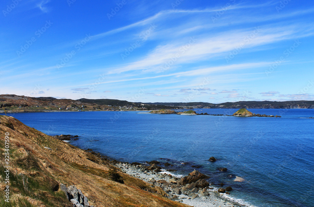 Looking along the cliffs and the coastline at the Atlantic Ocean, and the community of Ferryland, Newfoundland. Lots of blue sea, and blue sky with light, wispy clouds. 