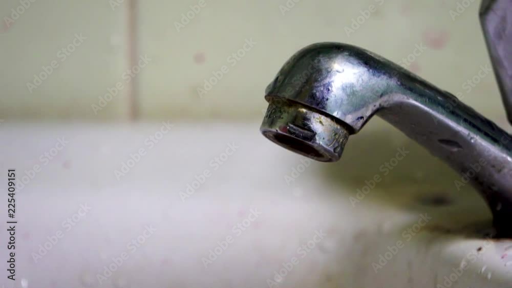 Dirty metal faucet slowly dripping droplets of water in a dirty sink with retro style green tile wall behind Dirty metal faucet slowly dripping droplets of water in a dirty sink with retro style green tile wall behind