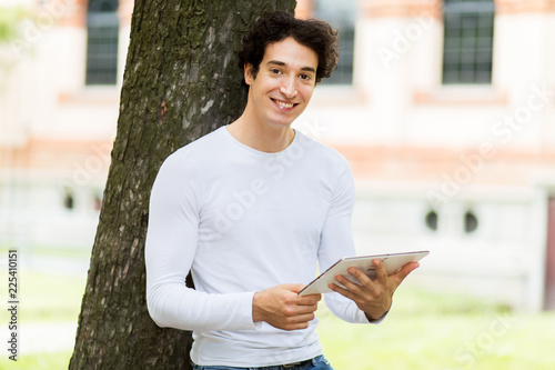 Young guy using a digital tablet lying against a tree in the park and enjoying the freedom of rhe wireless internet connection