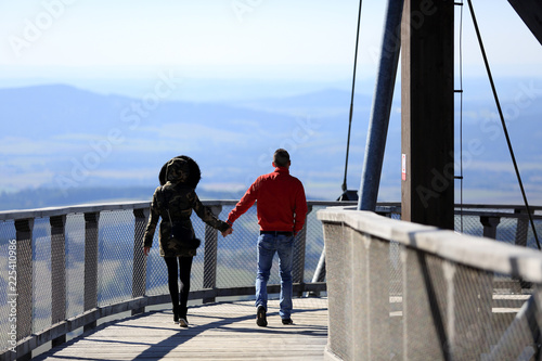 Kobieta i mężczyzna trzyma się za ręce idąc po platformie widokowej w górach.
