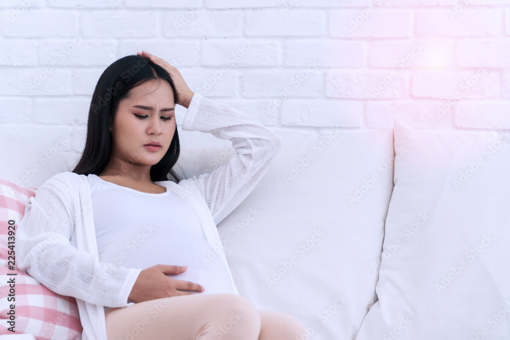 Pregnant young woman with hand on forehead suffering headache and abdominal pain sitting on white sofa in the living room at home. Pregnant young woman with hand on forehead suffering headache and abdominal pain sitting on white sofa in the living room at home.