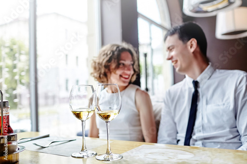 Two glasses of white wine standing on a wooden table in a restaurant. In the background a laughing couple in love. Selective focus