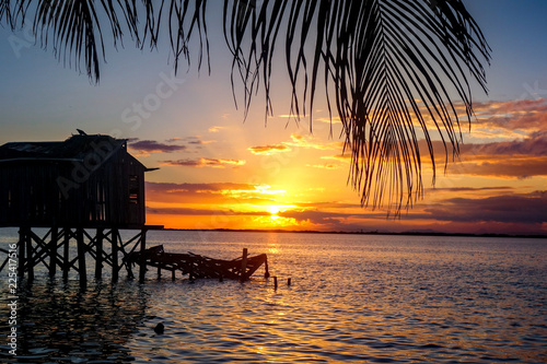An overwater shack is silhouetted by a dramatic orange sunset and framed by palm fronds on the tiny Caribbean Island of Tobacco Caye in Belize's barrier reef