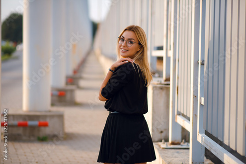 Smiling woman with blonde hair in glasses and black dress posing outdoors near the white columns. Urban background