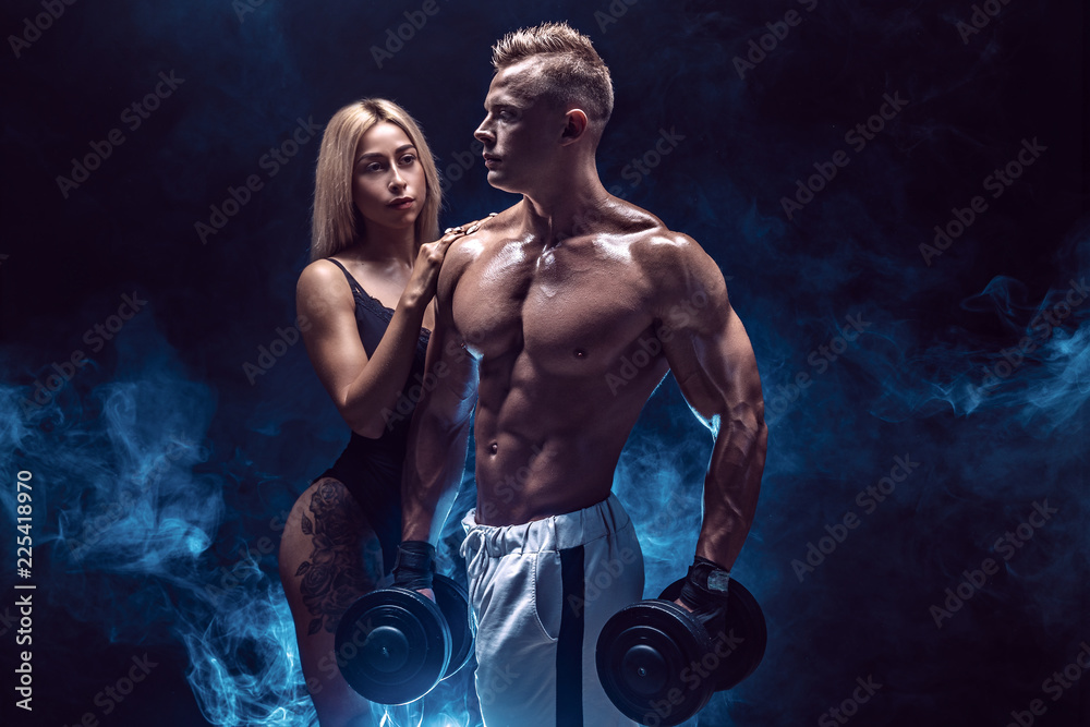 Attractive couple, a slim blonde female and handsome shirtless guy holding dumbbells, posing studio on a dark textured background with smoke.
