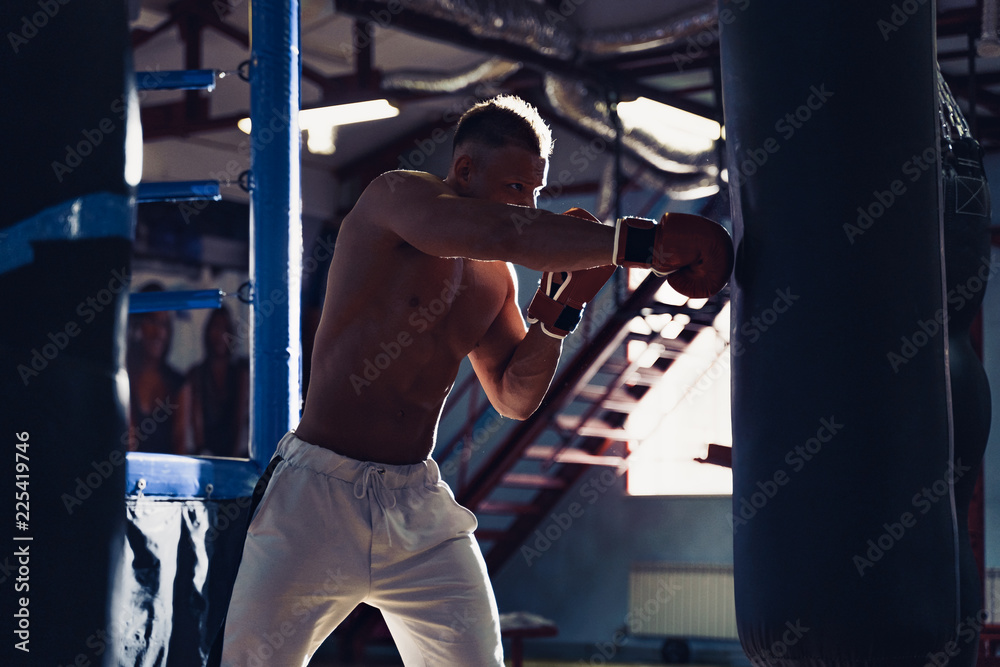 Male boxer training with punching bag in dark sports hall. Young boxer training on punching bag. Male boxer as exercise for the big fight. Boxer hits punching bag.