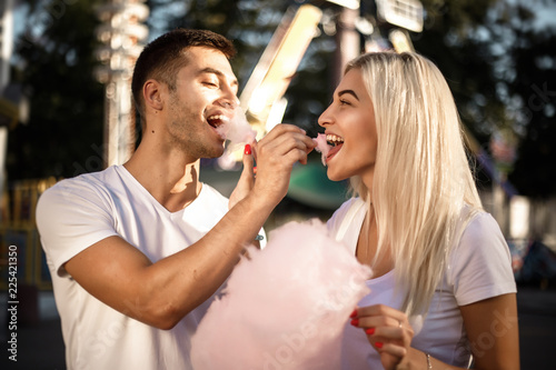 Happy young cheerful loving couple walking outdoors in the amusement park posing eat sweeties cotton candy floss.