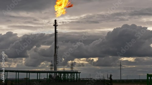 A tall GAS TORCH emits a bright flame into a cloudy evening sky at an oil facility. Industrial scene with heavy atmosphere and dramatic contrast.