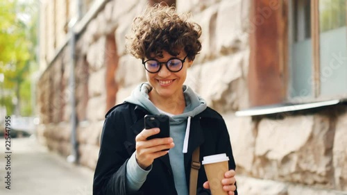 Smiling young woman in glasses is using smartphone looking at screen while walking outdoors in city with to-go coffee. Youth lifestyle, street and technology concept.