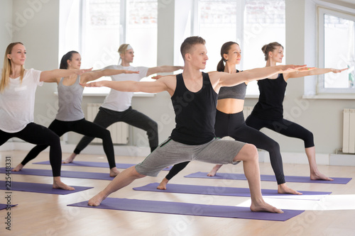 Group of young attractive sporty people practicing yoga lesson  doing Warrior Two exercise  Virabhadrasana II pose  working out indoor full length  students training in club studio. Well-being concept