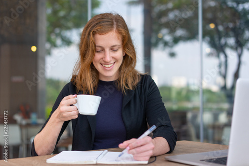 Positive student reading copybook during coffee break. Young woman in dark blue jacket drinking coffee and writing notes in notebook. Studying anywhere concept