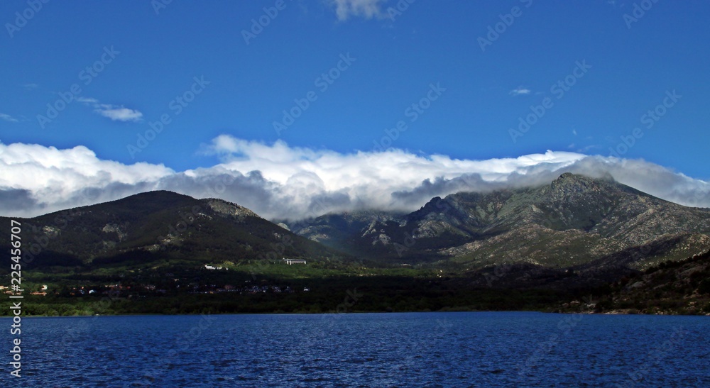 Panorámica de lago, bosques, montañas nevadas, nubes y cielo azul.