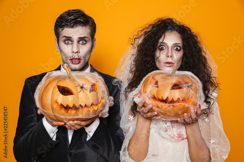 Surprised halloween bride and groom looking camera and holding pumpkin