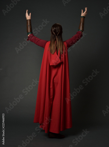 full length portrait of brunette girl wearing red medieval costume and cloak. standing pose with back to the camera on grey studio background.