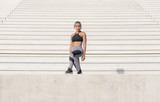 Middle Eastern Girl with short braided hair standing on the stairs of a construction site wearing gray and black fitness outfit on a hot bright sunny day.   