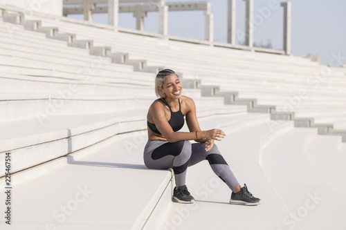Middle Eastern Girl with short braided hair sitting on the stairs laughing on a construction site wearing gray and black fitness outfit on a hot bright sunny day. 