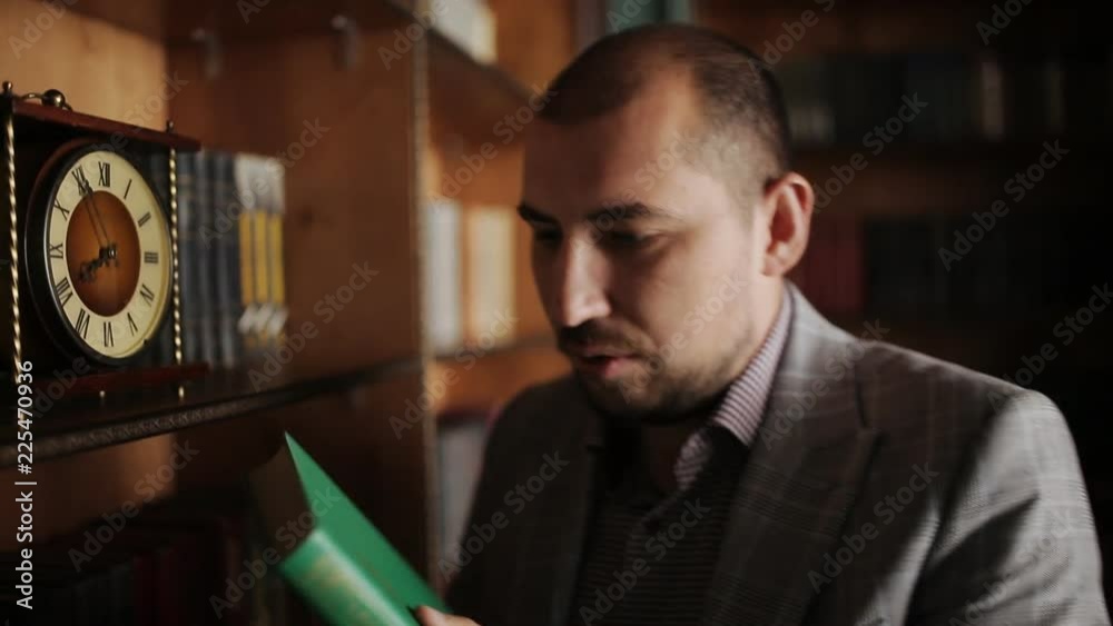 Mature professor is reading book standing in antique library, bearded man wearing eyeglasses is holding red exhibit, looking, having time on background of bookshelves in vintage room. Concept Mature professor is reading book standing in antique library, bearded man wearing eyeglasses is holding red exhibit, looking, having time on background of bookshelves in vintage room. Concept