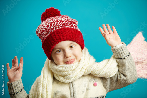 sweet little girl in winter hat and scarf, raised hands up, head leaned sideways, has a cute and charming face, stands on blue background.