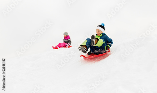 childhood, sledging and season concept - happy little kids sliding on sleds down snow hill in winter