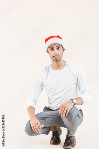shocked young man in santa hat crouching and looking at camera isolated on beige