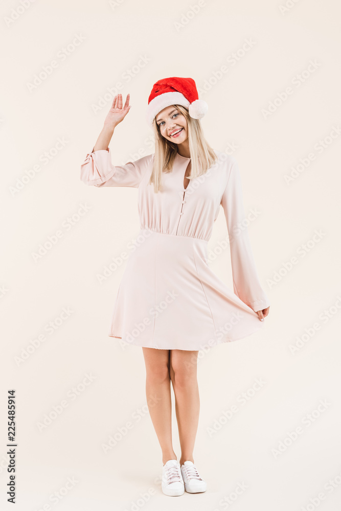 full length view of beautiful happy young woman in dress and santa hat smiling at camera isolated on beige full length view of beautiful happy young woman in dress and santa hat smiling at camera isolated on beige