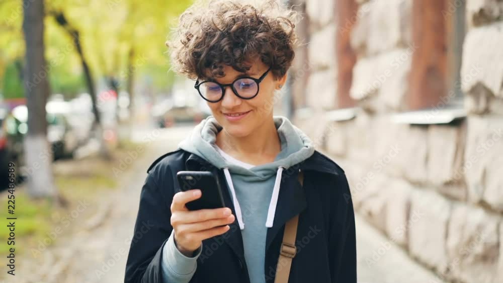 Slow motion of smiling woman in glasses and trendy clothing using smartphone looking at screen and walking outdoors in city. Youth lifestyle, street and technology concept.