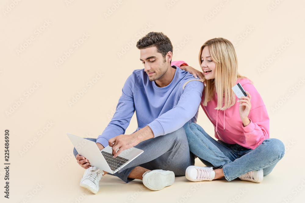 smiling young couple with credit card using laptop isolated on beige