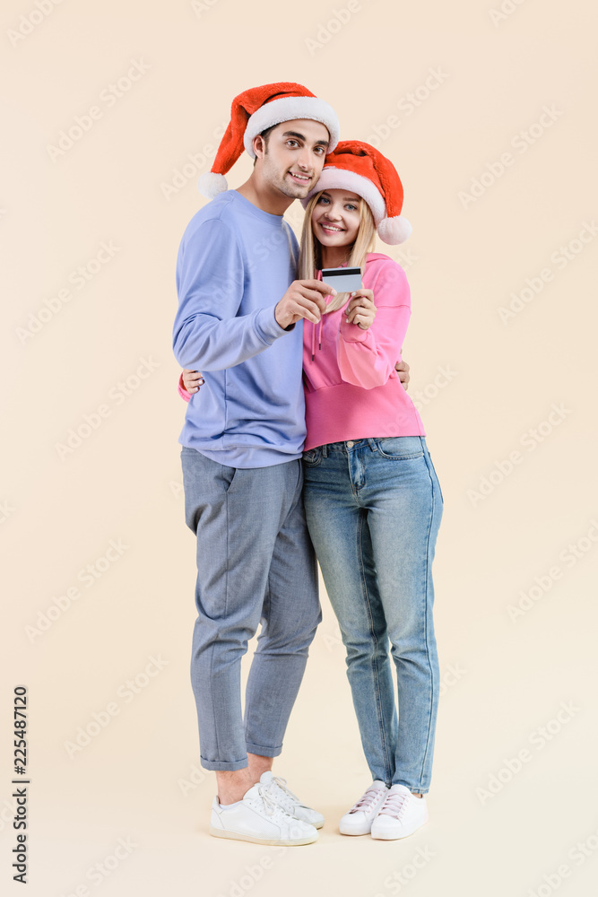 Obraz premium happy young couple in santa hats holding credit card and smiling at camera isolated on beige