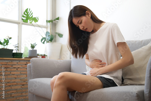 Asian girl having period is sitting on sofa and feeling much of painful on her stomach with unknown reason. She holds onto her stomach and curls through spasms. Healthcare and medical concept.