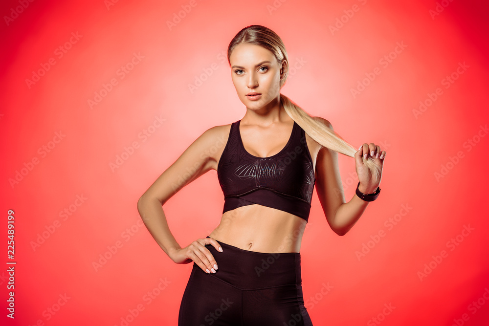 attractive sportswoman in black sportswear holding ponytail isolated on red and looking at camera attractive sportswoman in black sportswear holding ponytail isolated on red and looking at camera