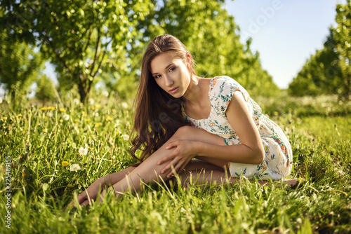 Beautiful model in a sitting position dressed in a flower dress, touch her bare legs. Rows of fruit trees in the background.