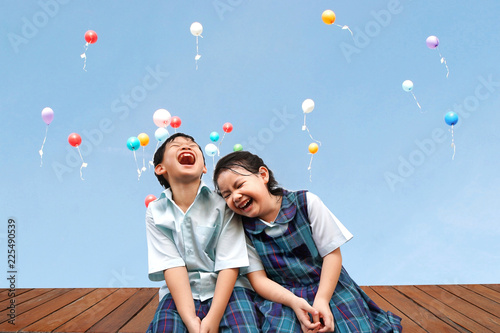 Funny Asian little kids brother and sister sitting on the wooden terrace with colourful balloons background.laughing and Happiness of cute kid in uniform school,Education,Family,Children Concept.