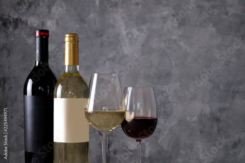 Two bottles of red and white wine with matte black & white blank labels on concrete wall background. Wineglasses half full with vintage expensive wine of different type. Close up, copy space.