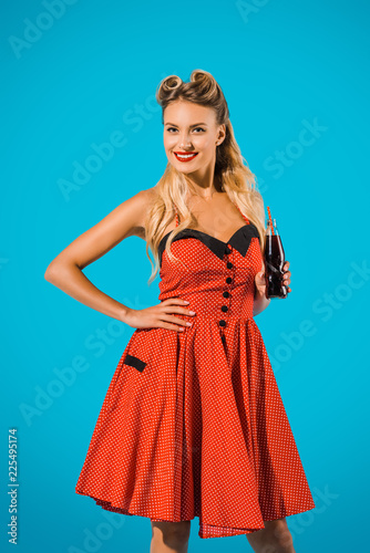 portrait of smiling pin up woman in vintage dress with soda drink in glass bottle on blue background